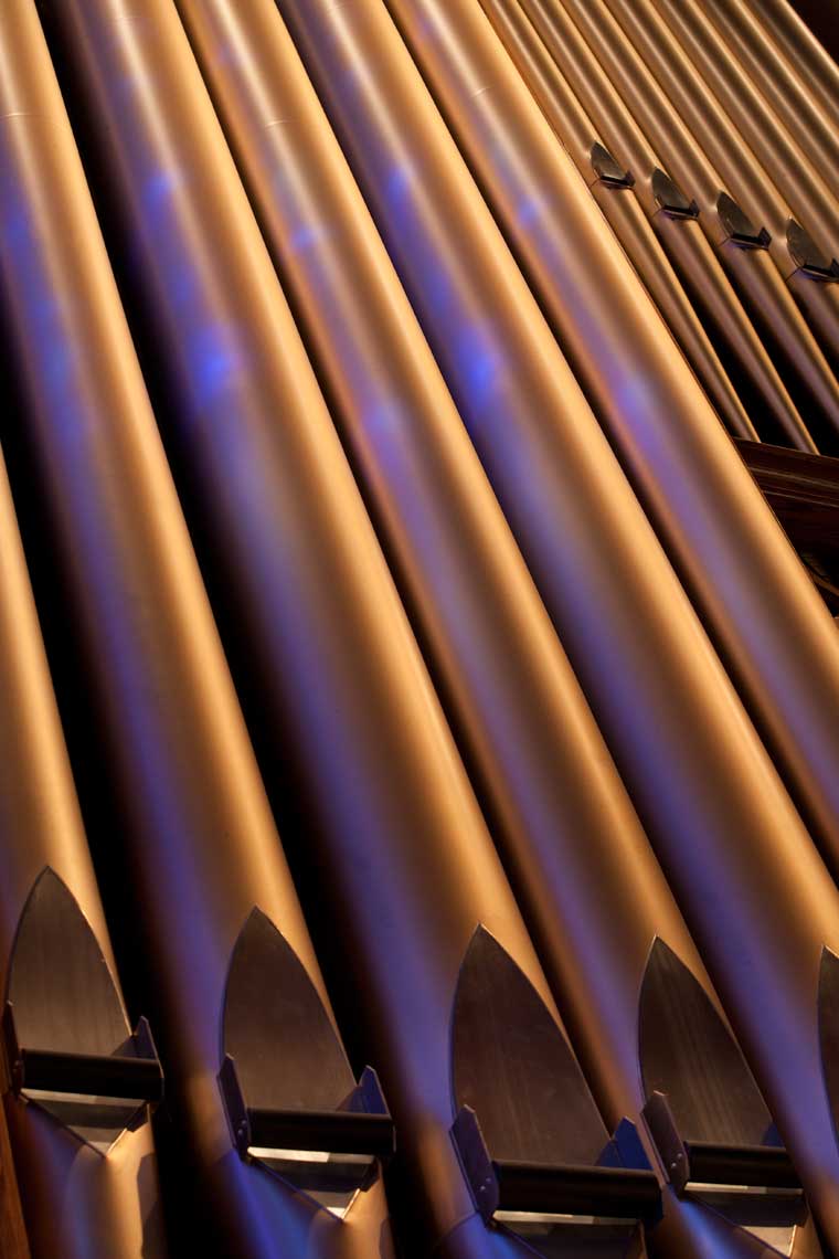 A detail of pipe organ pipes, with light from the stained-glass window falling upon them