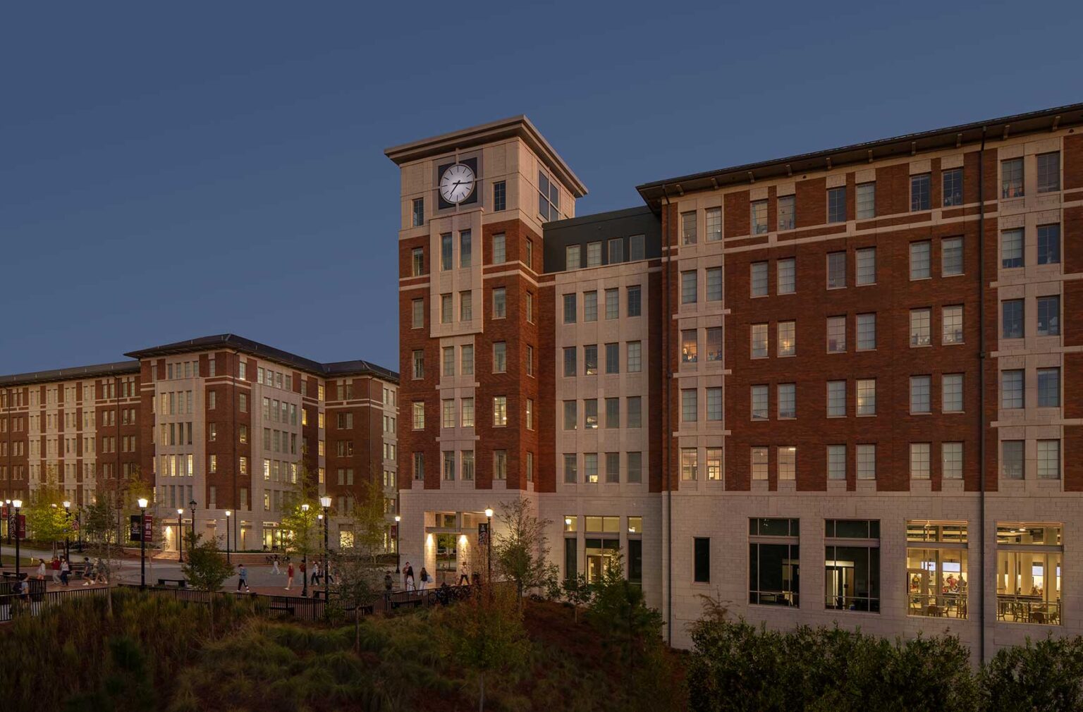 A twilight view of the clock tower and plaza at the University of South Carolina Campus Village with students enjoying the campus.