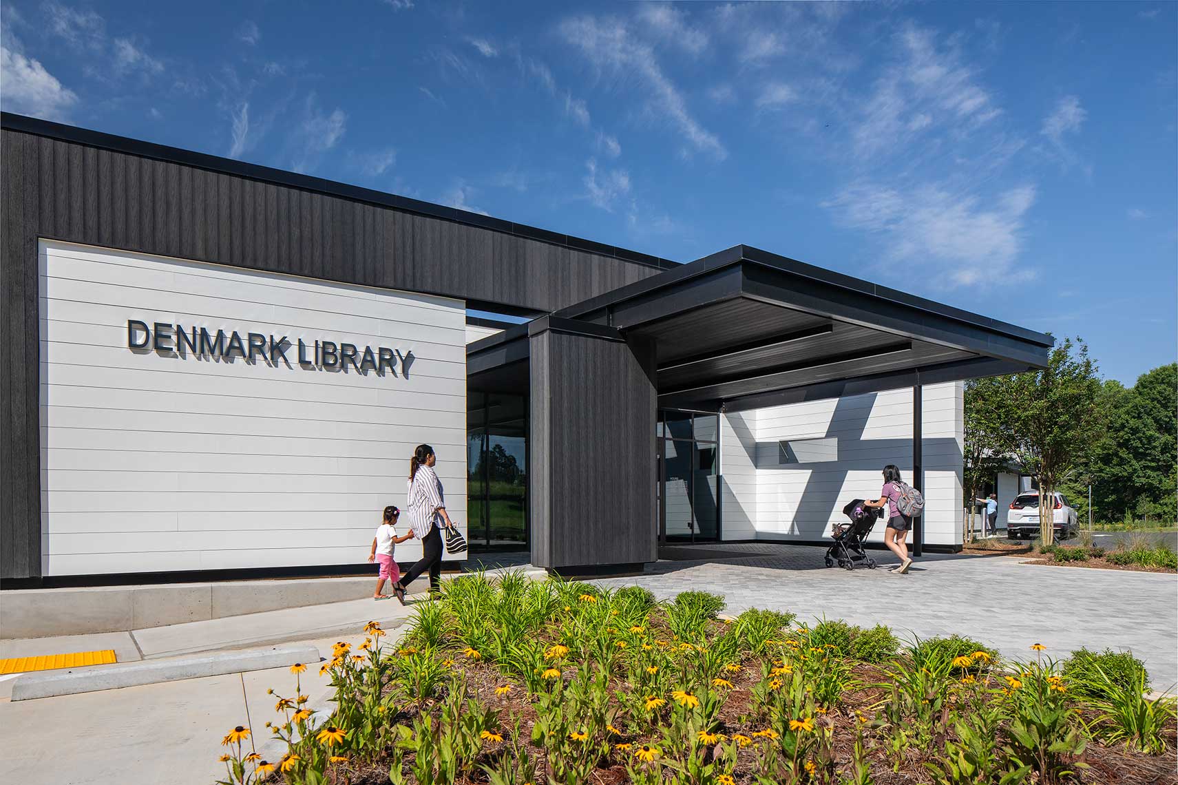 Library patrons stroll into the inviting main entry of the Denmark Branch Library, and one patron returns books from her car in the drive-up lane.