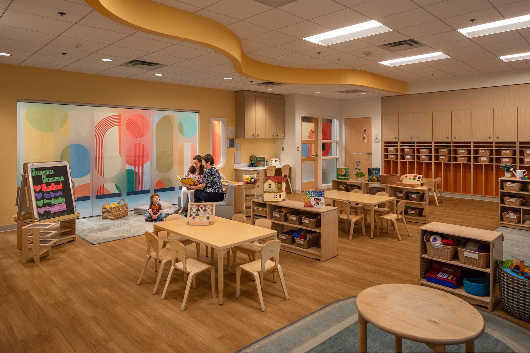 An interior view of a colorful and whimsically designed Preschool classroom, with two teachers reading to a student
